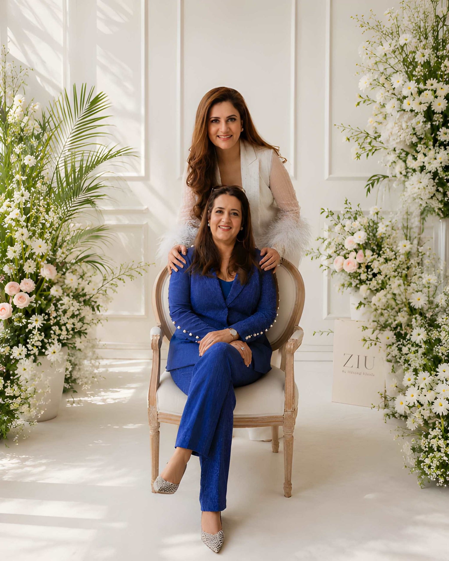 Two women (Pooja khosla & Shivangi Khosla) posing together in a decorated room with flowers and a chair.